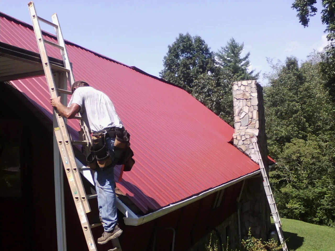 Roofer on a ladder beside a red metal roof and stone chimney — Soffit & Fascia Repair by Roofing Brothers in Cottondale